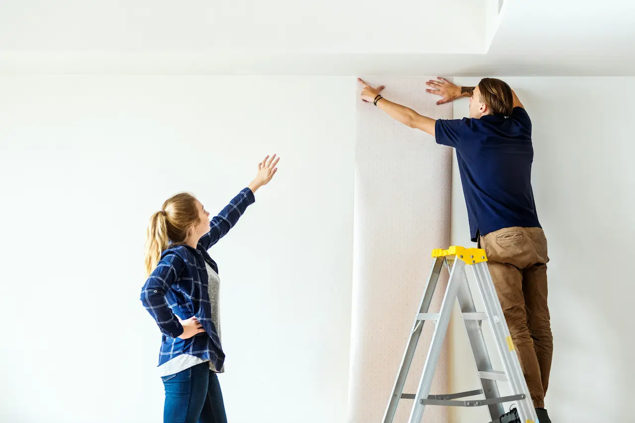 Personne sur un escabeau posant un lé au haut d'un mur pendant qu'une autre guide l'alignement
